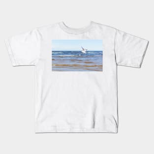 White seagull flying above the water to the left with its wings open Kids T-Shirt